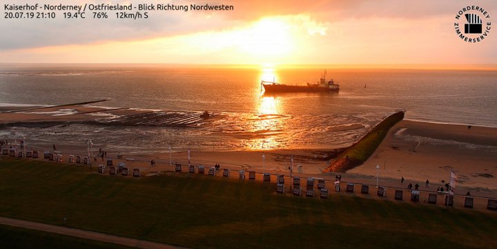 Sonnenuntergang über der Nordsee am 20. Juli 2019: Auf der Nordsee fährt ein Schiff, im Vordergrund die Strandpromenade von Norderney mit Strandkörben und Fahnen und Menschen